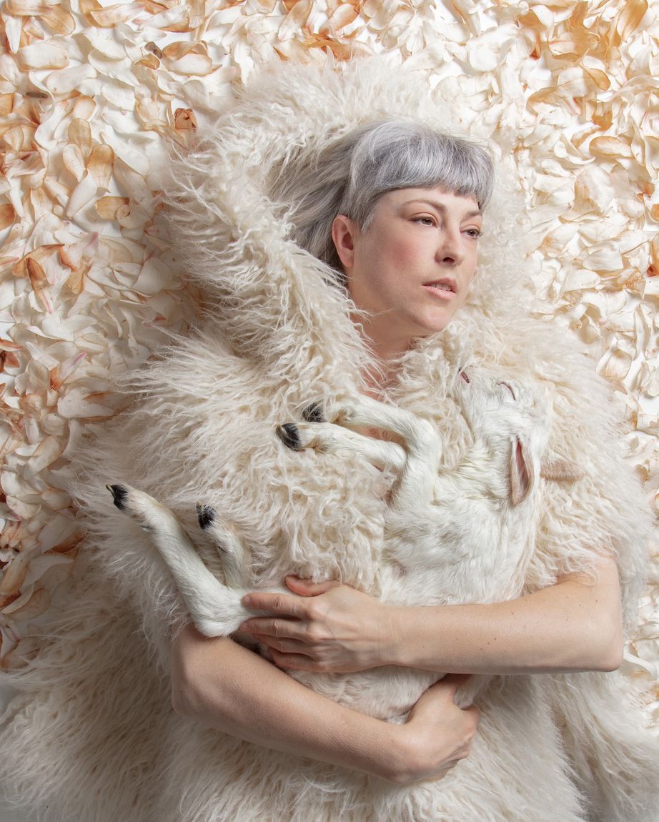 The artist who has a short gray bob haircut is wearing a fuzzy white lamb fur cape and holding a white still-born lamb in her arms. They are lying on a bed of white magnolia petals. 