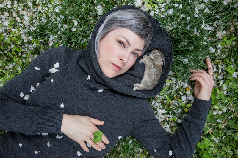 The artist is reclining in grass that is full of white crab apple petals. Petals fall across her gray cowl neck sweater which has been pulled up and around her face to create a bowl, and a perfect, dead baby cottontail rabbit is nestled into the sweater near her face. A four leaf clover is held in her fingers across her chest.
