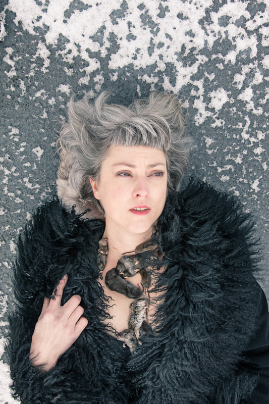 The female artist who has a short gray bob haircut lies on gray, snowy cement. She is wearing a black fur coat and several dead rodents lie across her bare chest and throat. 